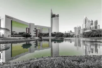 View of the buildings in Dubai Media City (DMC), including the large Oracle building, reflected in a still lake with surrounding greenery. The image is black and white with a green tint on the sky and reflections.