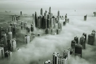 Atmospheric aerial view of a dense cluster of Dubai skyscrapers emerging from a thick blanket of fog or clouds. The image is black and white, symbolizing the large scale of the Jebel Ali Free Zone (JAFZA) area.