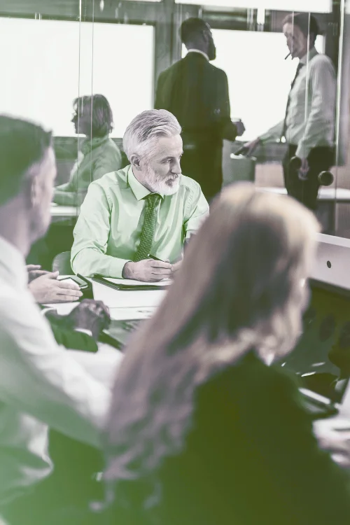 A business meeting in a bright, modern office. A smiling, grey-haired man in a prominent green shirt sits at the table, while other colleagues are reflected in the glass walls. The image represents the benefits of a Mainland Company setup.