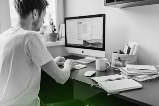 Man working from a home office, typing on a desktop computer keyboard with notebooks, a mouse, and a coffee mug on the desk.