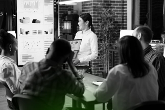 Businesswoman giving a presentation, pointing to a large screen displaying charts, graphs, and market data to her team in a meeting.