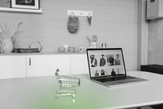 Black and white photo of a tiny shopping cart next to a laptop displaying an e-commerce clothing website on a table.