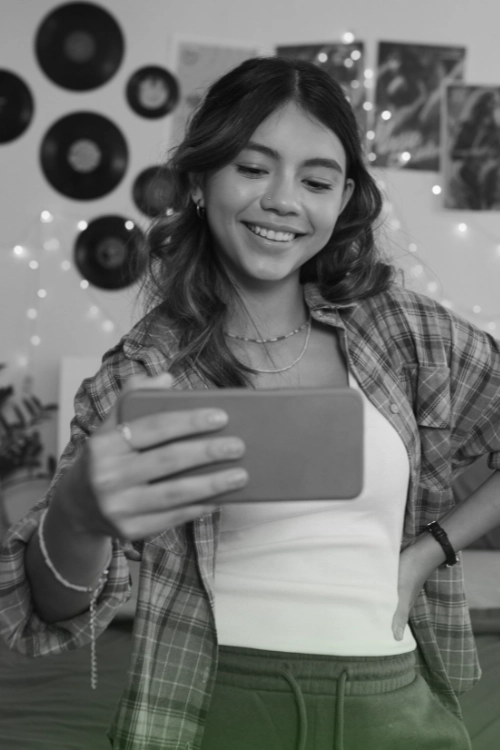 Smiling young woman holding a smartphone, taking a selfie, or creating social media content in a casual home setting.