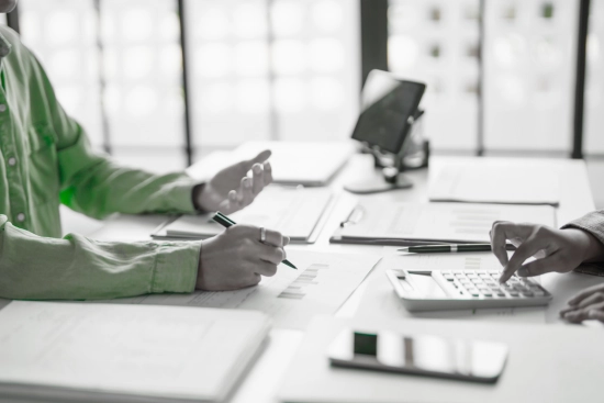 Two people in a business meeting, one writing on a document and the other operating a calculator, discussing financial reports or forecasts.