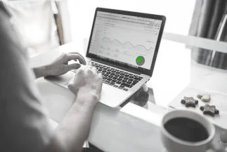A person's hands typing on a laptop displaying Google Ads or analytics performance graphs, with a cup of coffee and cookies nearby, symbolizing managing online advertising campaigns.
