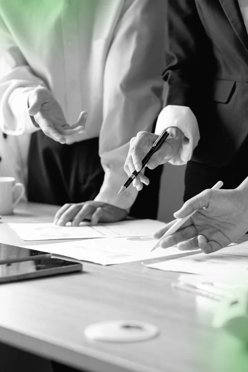 A close-up of three people's hands pointing at and discussing documents spread out on a table, with one person holding a pen, symbolizing a detailed business meeting or outlining a work process.