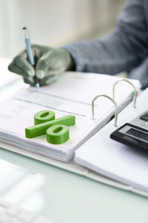 A person writing on documents in a binder, with a large, green percent sign (%) figurine on top of the paperwork, symbolizing tax preparation or calculating percentages.
