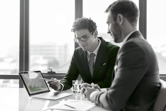 Two business professionals in suits reviewing a financial graph on a laptop screen during a meeting. The person on the left is pointing to the screen with a pen.