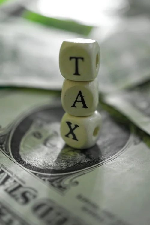 Close-up of three white wooden dice stacked on top of one another, spelling "T A X" (top to bottom), resting on top of US dollar bills.