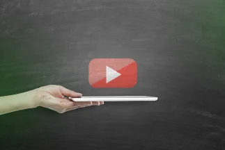 A hand holding a white tablet horizontally against a dark background, with a prominent floating YouTube play button icon above it, symbolizing video and YouTube advertising.