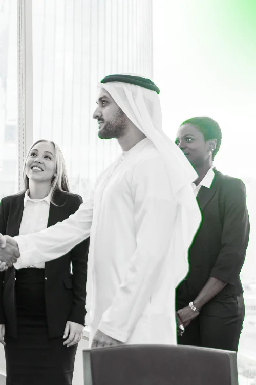 A diverse business team: a man in traditional Emirati kandura and headwear is shaking hands with a smiling woman in a suit, while another woman looks on. This image represents business partnership and success in the UAE.