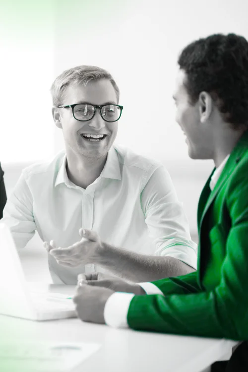 Two professional men smiling and engaged in discussion at a table with a laptop; one man in a white shirt and glasses is gesturing, and the other is wearing a bright green blazer, symbolizing a consultation or Q&A session.